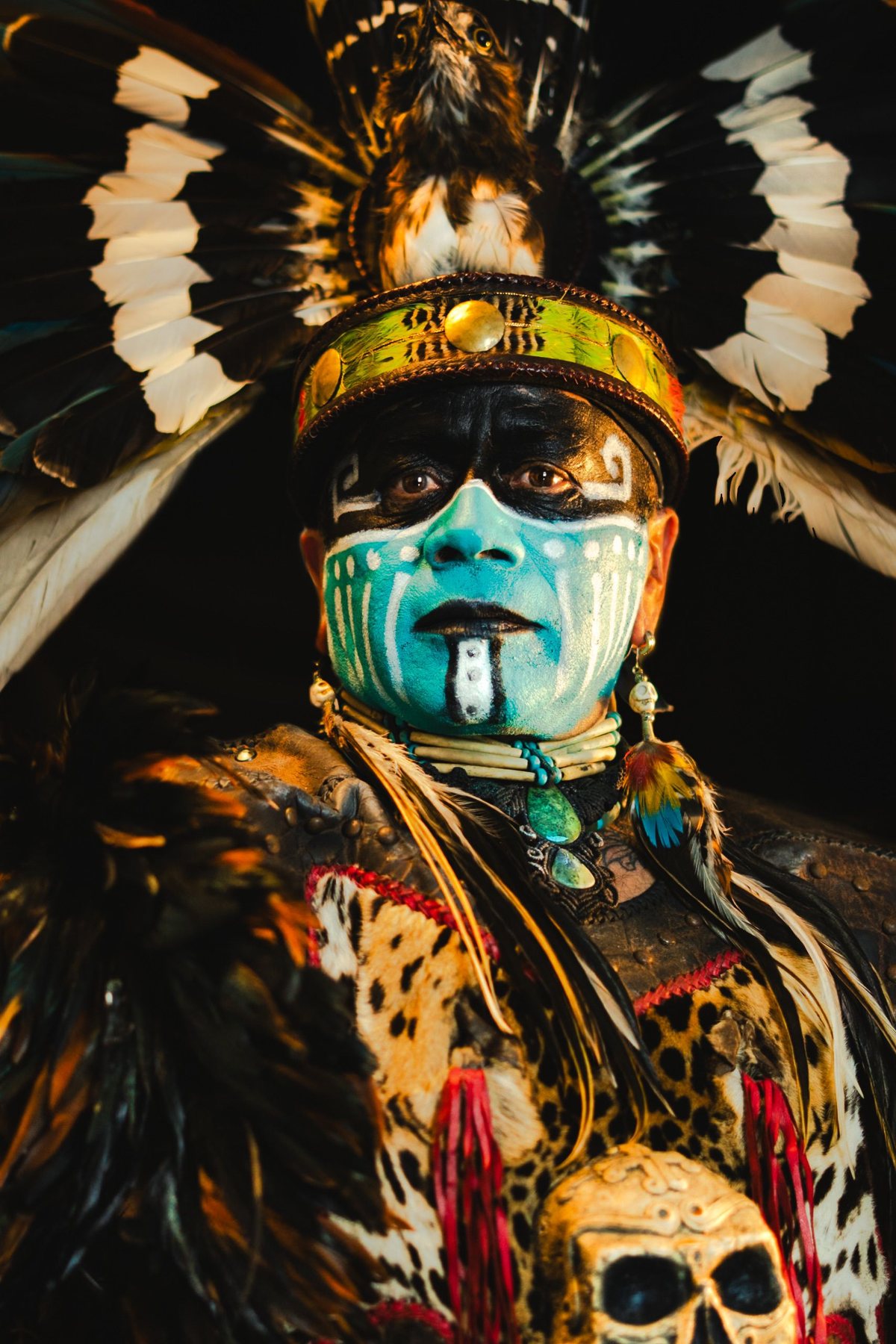 An elder in full ceremonial regalia — painted face, raptor headdress, jaguar mantle — gazing directly forward with absolute composure.
